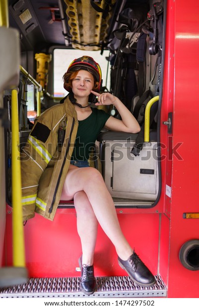 Image of ginger firewoman in helmet looking\
in camera sitting in cab of fire\
engine