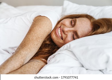 Image of ginger happy woman with freckles smiling at camera while lying in bed at home