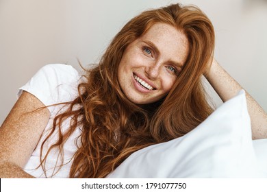 Image of ginger happy woman with freckles smiling at camera while lying in bed at home