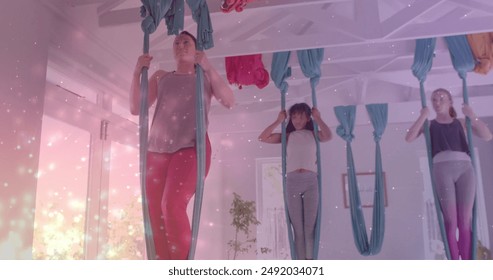 Image of glowing particles over diverse female instructor and teenagers practicing aerial yoga. Wellbeing, energy, healthy lifestyle, connection and communication, digitally generated image. - Powered by Shutterstock