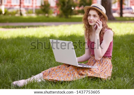 Image of good looking curly haired young female sitting on grass, wearing colorful skirt, red top and straw hat, relaxing in park, having rest during break, talking over phone, using laptop.
