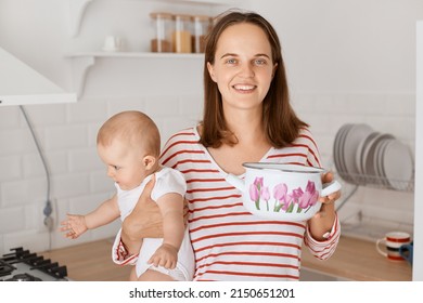 Image Of Good Looking Pretty Young Mother Posing With Toddler Daughter In Kitchen, Holding Pot With Soup, Looking At Camera With Positive Expression, Cooking With Child.