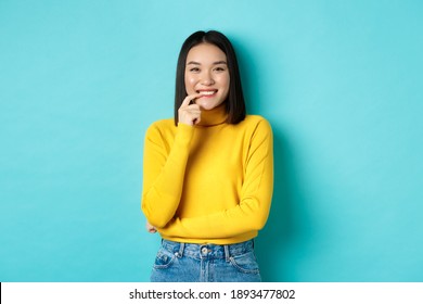 Image Of Good-looking Asian Woman In Stylish Outfit, Touching Lip And Smiling At Camera With Happy Face, Standing Over Blue Background