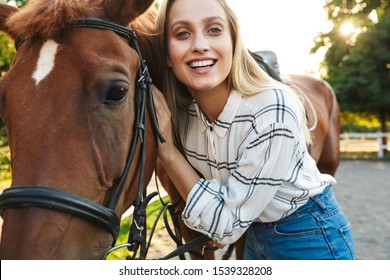 Image Of Gorgeous Young Blonde Woman Smiling And Standing By Horse At Yard In Countryside