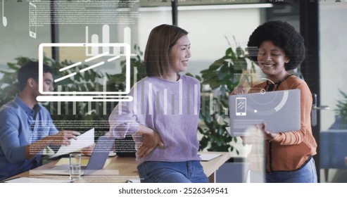 Image of graphs, icons and trading board over diverse coworkers discussing reports on laptop. Digital composite, multiple exposure, business, stock market, teamwork, planning and technology.