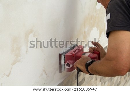 Image of the hands of a handyman who removes plaster and mold from a wall with a sanding machine. Reference to problems of humidity