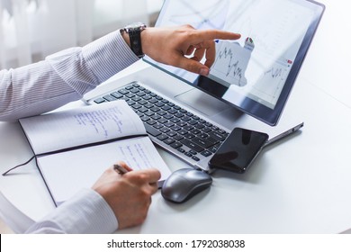 The Image Of The Hands Of A Young Man Sitting At A Table Writing In A Notepad With A Ballpoint Pen, The Second Hand Shows The Laptop Screen. On The Screen Of The Laptop Depicts Graphs