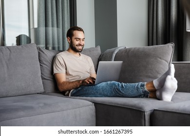 Image of a handsome cheerful young man indoors at home on sofa using laptop computer.