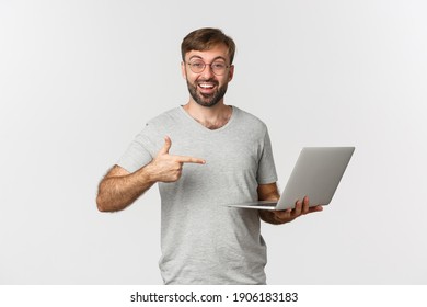 Image of handsome man with beard, wearing glasses and gray t-shirt, pointing at laptop and smiling, recommend something, standing over white background