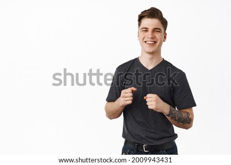 Image of handsome smiling guy awkward dancing. Young man laughing and holding fists clenched for good luck, standing against white background, holding in hands