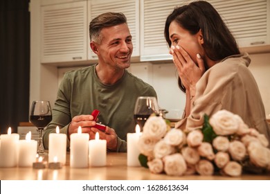 Image of happy adult man making proposal to his girlfriend with engagement ring during romantic candlelight dinner at home