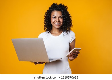 Image of happy african american woman holding cellphone and laptop isolated over yellow background