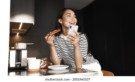 Image of happy asian woman eating breakfast and using smartphone. Girl laughing, eat toast and drink coffee, holding mobile phone in kitchen at home - Shutterstock ID 2001960107