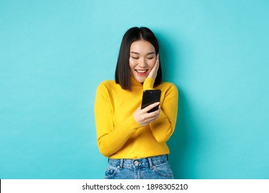 Image Of Happy Asian Woman Reading Message On Mobile Phone Screen And Smiling, Chat In Smartphone App, Standing Over Blue Background