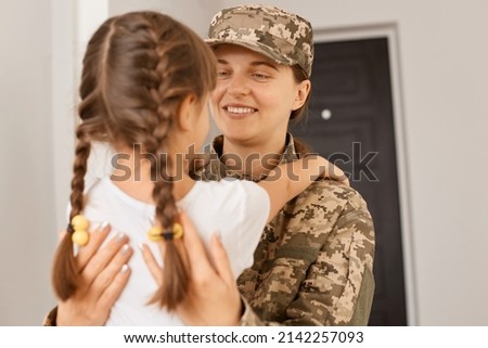 Image of happy attractive military woman wearing camouflage uniform and cap posing with her daughter, people hugging each other, mom looking with gentle at her child.