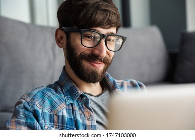 Image of happy bearded man in eyeglasses smiling and using notebook while sitting in living room - Shutterstock ID 1747103000