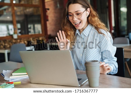 Image of happy beautiful woman waving hand and drinking coffee takeaway while using laptop at classroom
