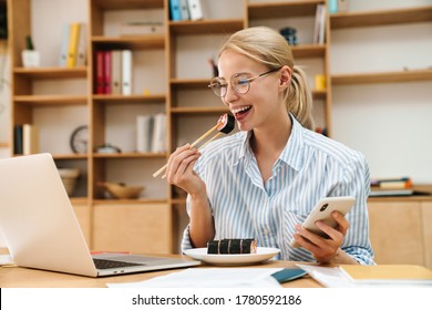 Image of happy blonde woman eating sushi with chopsticks while using cellphone and laptop in office - Powered by Shutterstock