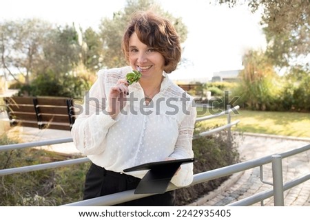 Image of happy caucasian business woman 45 years old, company worker, smiling and holding digital tablet, standing outside, holding park flower near her lips. Concept of women's health, allergy, break