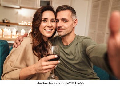Image of happy caucasian couple taking selfie photo while having romantic candlelight dinner at home