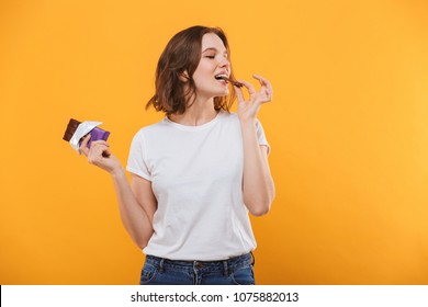 Image of happy cute young woman standing isolated over yellow background eating chocolate.