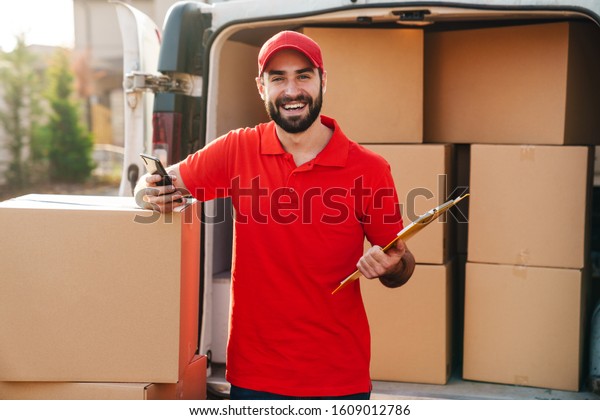 Image of happy\
delivery man holding clipboard and using cellphone while standing\
with parcel boxes\
outdoors
