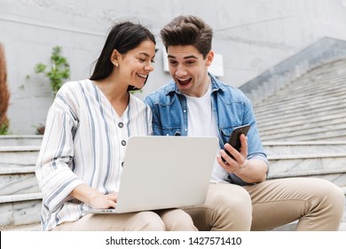 Image Of Happy Excited Shocked Young Amazing Loving Couple Business People Colleagues Outdoors Outside On Steps Using Mobile Phone And Laptop Computer.