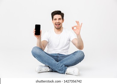 Image of a happy excited young man posing isolated over white wall background using mobile phone showing display sitting on floor make okay gesture.