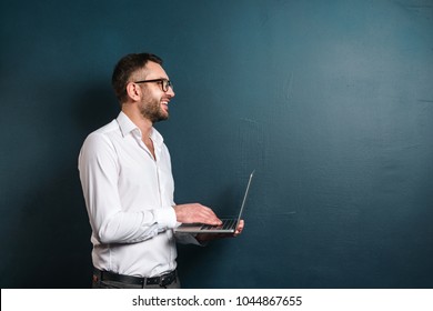 Image of happy man wearing glasses standing over dark blue background looking aside using laptop computer.