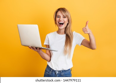 Image Of A Happy Optimistic Emotional Young Blonde Woman Posing Isolated Over Yellow Wall Background Dressed In White Casual T-shirt Using Laptop Computer Showing Thumbs Up Gesture.