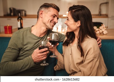 Image of happy passionate couple drinking red wine from glasses while having romantic candlelight dinner at home
