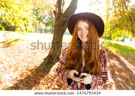 Image of a happy positive cutie young student redhead girl in autumn park using mobile phone.