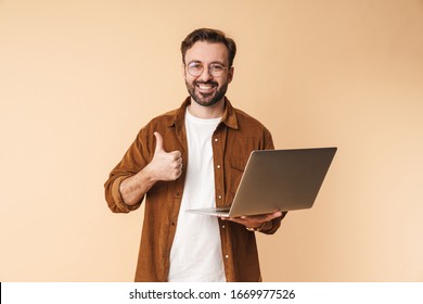 Image of a happy positive young unshaved man isolated over beige wall background using laptop computer showing thumbs up. - Shutterstock ID 1669977526
