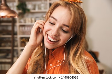 Image of a happy smiling cute young blonde girl in orange shirt at the kitchen listening music with earphones. - Shutterstock ID 1440904016