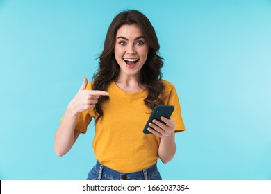 Image of a happy surprised young girl isolated over blue wall background using mobile phone pointing.