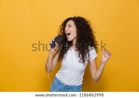 Image of happy woman 20s with curly hair singing while holding smartphone like microphone and listening to music via earphones isolated over yellow background