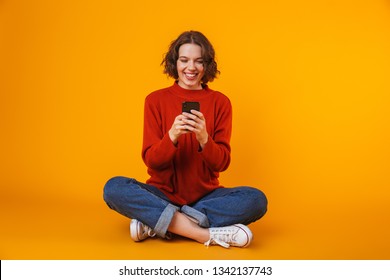 Image of happy woman 20s wearing sweater using smartphone and smiling while sitting on the floor isolated over yellow background
