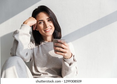 Image of happy woman 30s holding cup with tea while sitting over white wall indoor