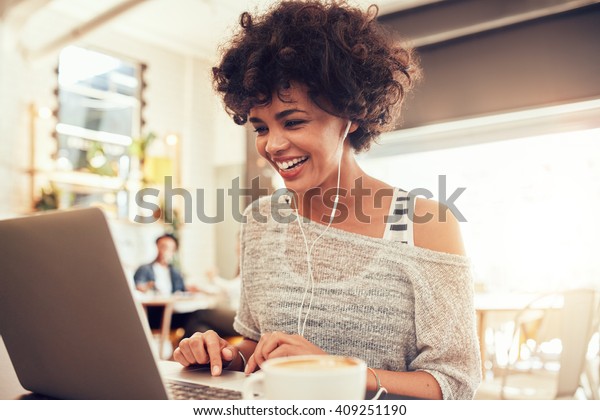 Image of happy woman using laptop while sitting\
at cafe. Young african american woman sitting in a coffee shop and\
working on laptop.
