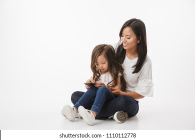 Image Of Happy Young Asian Woman Mother With Her Little Girl Child Daughter Sitting Isolated Over White Wall Background Using Mobile Phone.