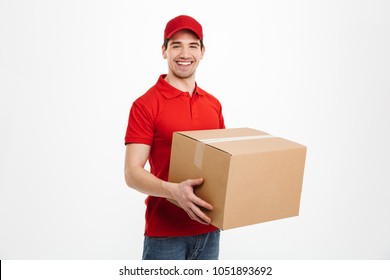 Image of a happy young delivery man in red cap standing with parcel post box isolated over white background.