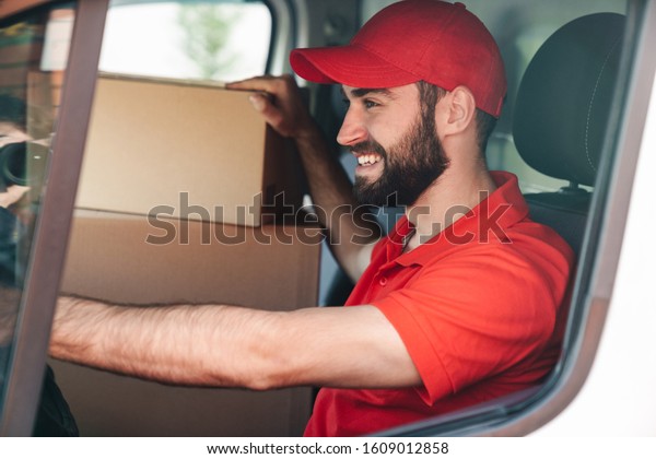 Image of happy young delivery man in\
red uniform smiling and driving van with parcel\
boxes