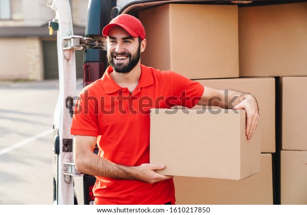 Image of happy young delivery\
man in red uniform standing with parcel post box near car\
outdoors