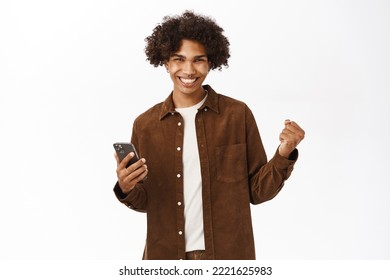 Image Of Happy Young Hispanic Boy Holding Mobile Phone And Triumphing, Celebrating Good News, Standing Over White Background