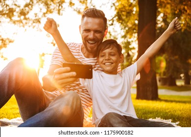 Image of happy young man father have a rest with his son outdoors in park play games with mobile phone. - Powered by Shutterstock