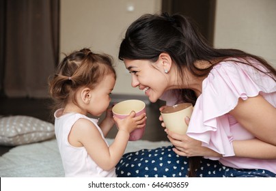 Image of happy young mother drinking tea with her little daughter sitting on bed indoors. Looking aside. స్టాక్ ఫోటో