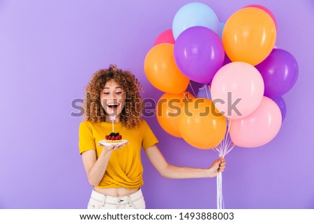 Image of happy young woman celebrating birthday with multicolored air balloons and piece of pie isolated over violet background