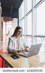 Image Of A Happy Young Woman In A Jacket Smiling And Working On A Laptop While Talking On The Phone In A Modern Office With Large Windows. Remote Work