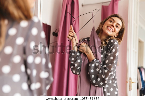 Image of happy young woman shopaholic\
standing near changing room indoors looking at\
mirror.