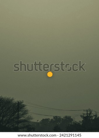 Image of a hazy, overcast sky with the sun appearing as a bright, distinct circle amidst the grey atmosphere, with silhouettes of trees and utility poles.